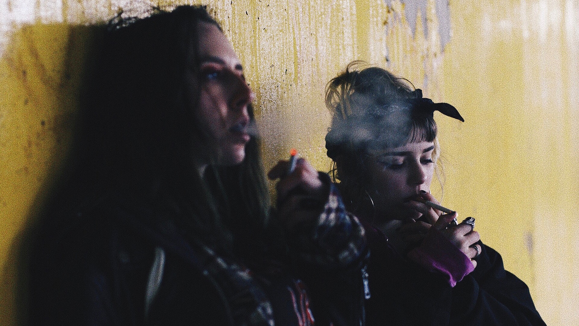 two teen girls smoking self harm up against a yellow wall